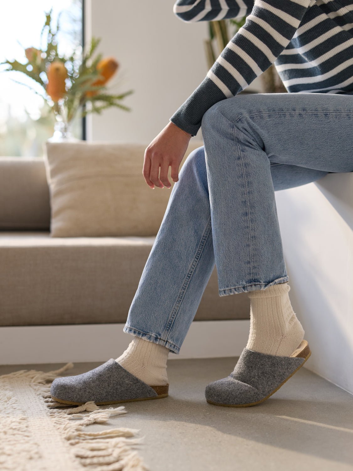 A person in a striped sweater, light jeans, and Cozy Earth's Lakehouse Clog sits on a white bench with one foot crossed over the knee. A beige rug is underfoot, and flowers adorn the bright window in the background. |Color:Charcoal