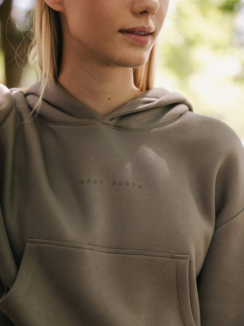 A person wearing a Women's CityScape Cropped Hoodie in olive-green, embellished with the subtle text "Cozy Earth," stands in soft, natural light. The background is blurred, featuring greenery that suggests an outdoor setting. The photo is cropped at the shoulders, with only part of the person's face visible.