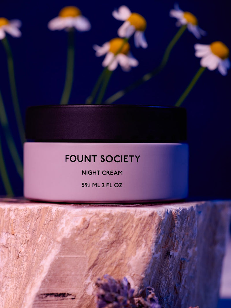 A jar of Cozy Earth Night Cream sits on a wooden surface, its label clearly visible. In the backdrop, soft-focus white daisies with yellow centers stand out against a blue background.