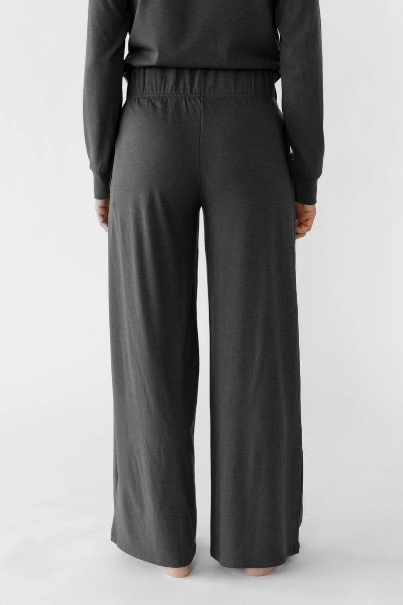A person stands barefoot against a light background, wearing Cozy Earth's Women's Ultra-Soft Bamboo Wide Leg Pull On Pant & Pullover Set, featuring a dark, loose-fitting long-sleeved top and wide-leg pants.