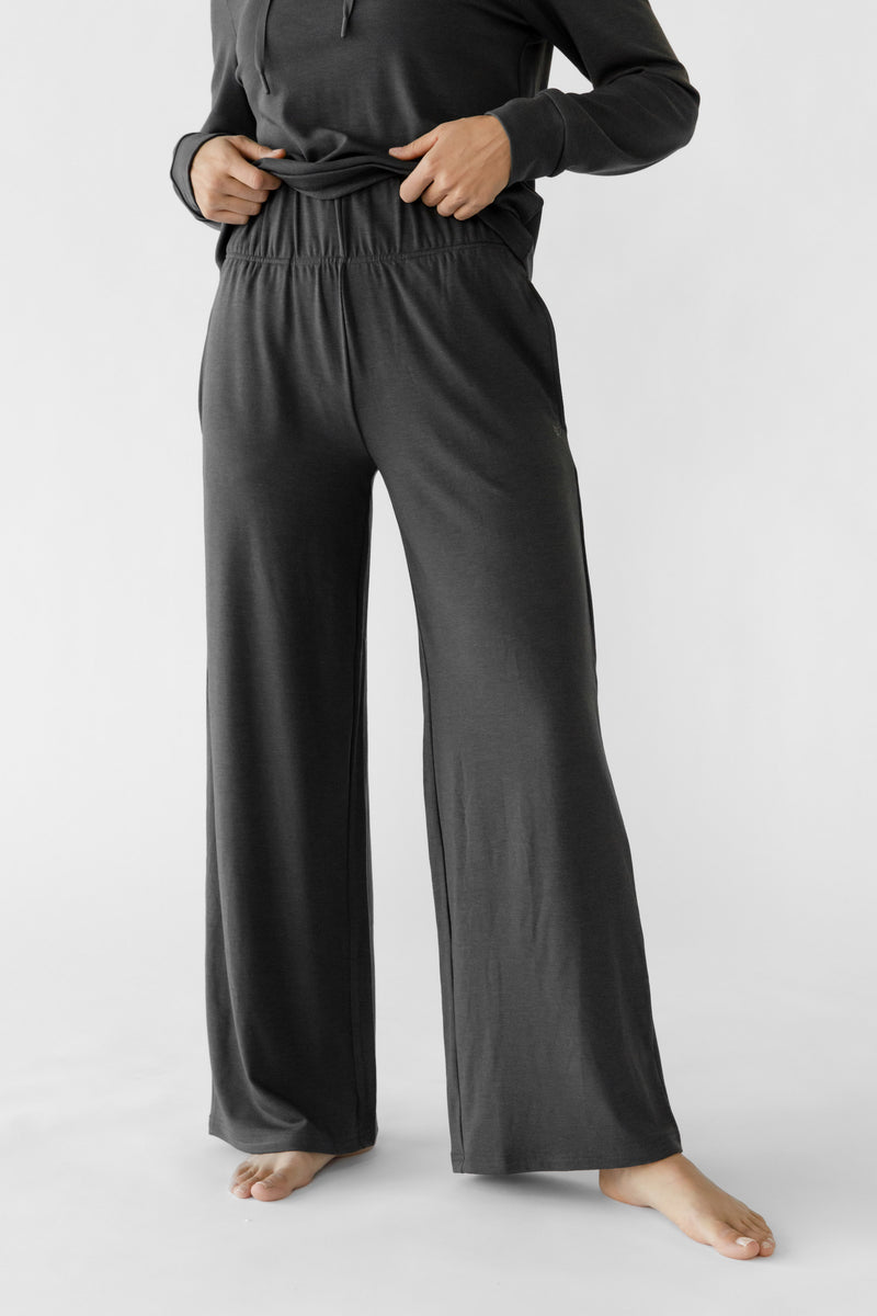A person dressed in Cozy Earth's Women's Ultra-Soft Bamboo Wide Leg Pull On Pant & Pullover Set in dark gray adjusts the waistband while barefoot against a light background.