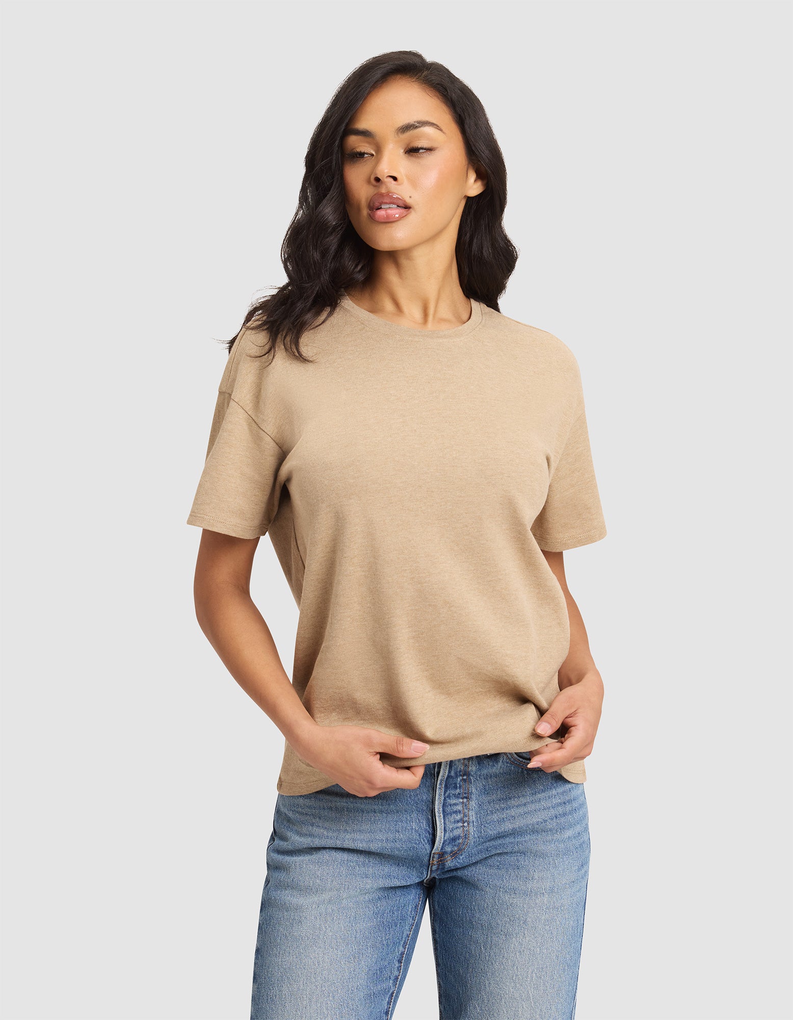 A woman with long dark hair models the Cozy Earth Women's All Day Tee in beige, paired with blue jeans, against a plain light gray background. She looks slightly to the side and holds the hem of her shirt with one hand. 