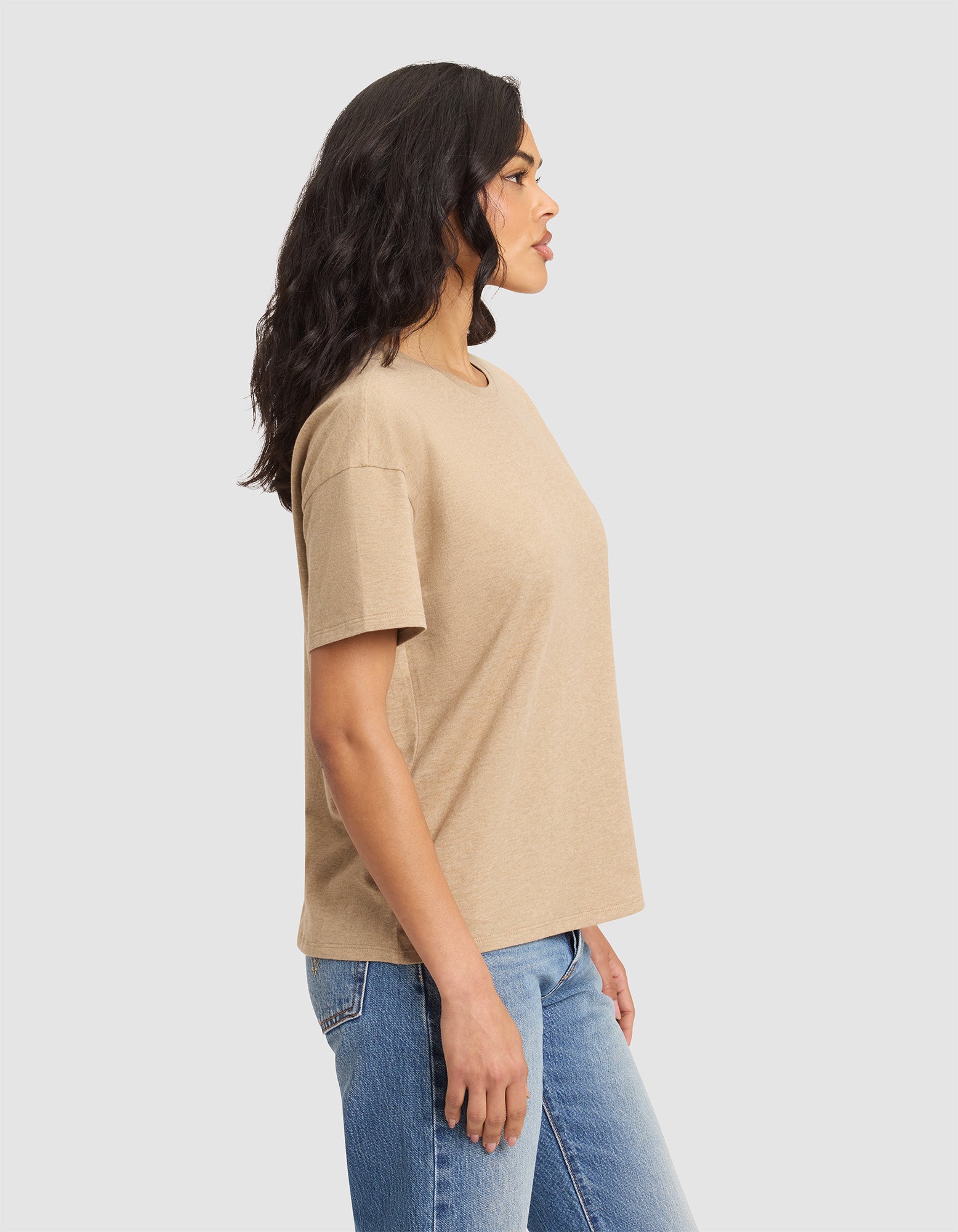A woman with wavy dark hair stands in profile, wearing Cozy Earth's Women's All Day Tee in beige and blue jeans, set against a plain light gray background. 