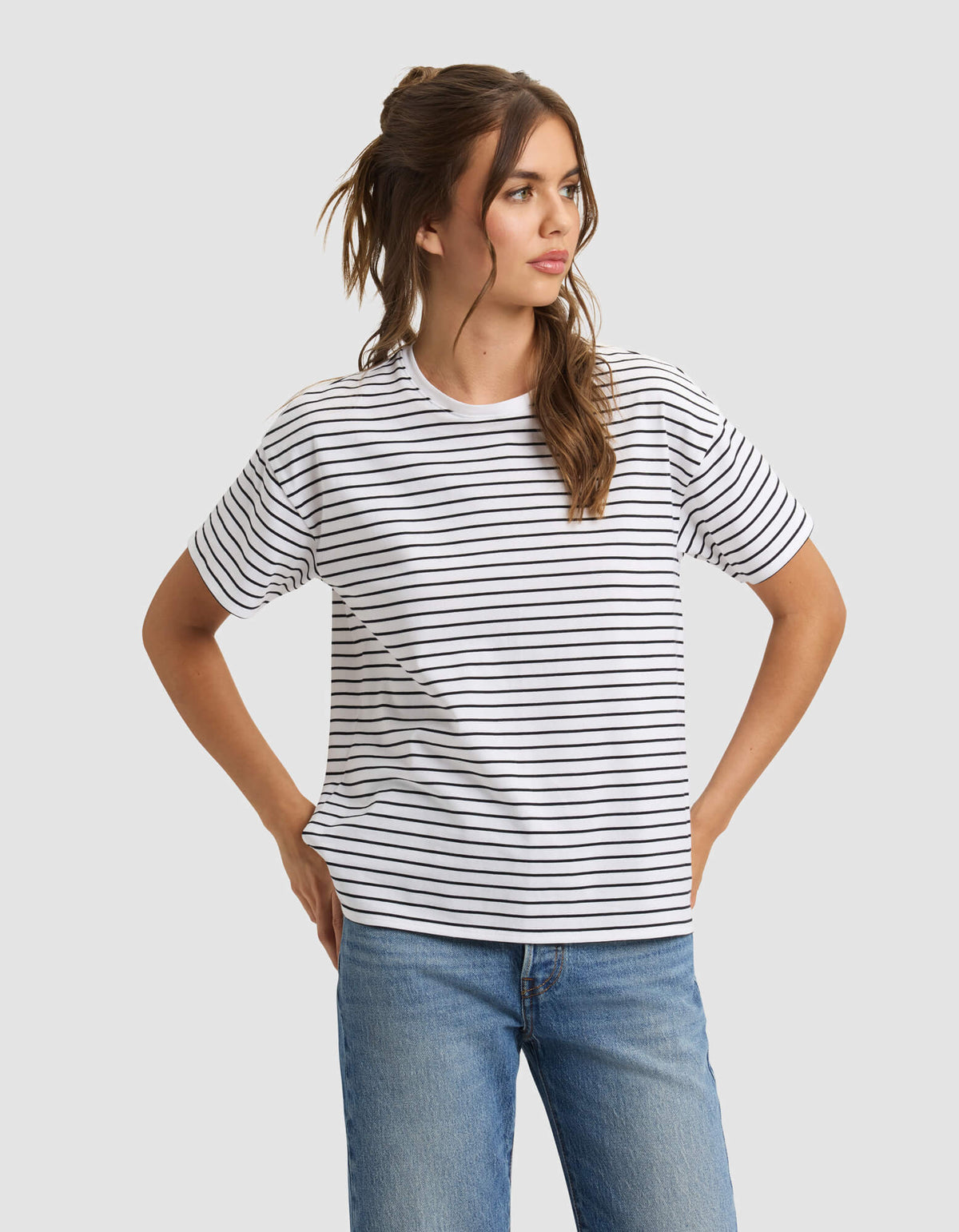 A woman models the Cozy Earth Women's All Day Tee—white with black horizontal stripes—paired with blue jeans, standing hands on hips and looking to the side against a plain light gray background. |Color:Jet Black Stripe