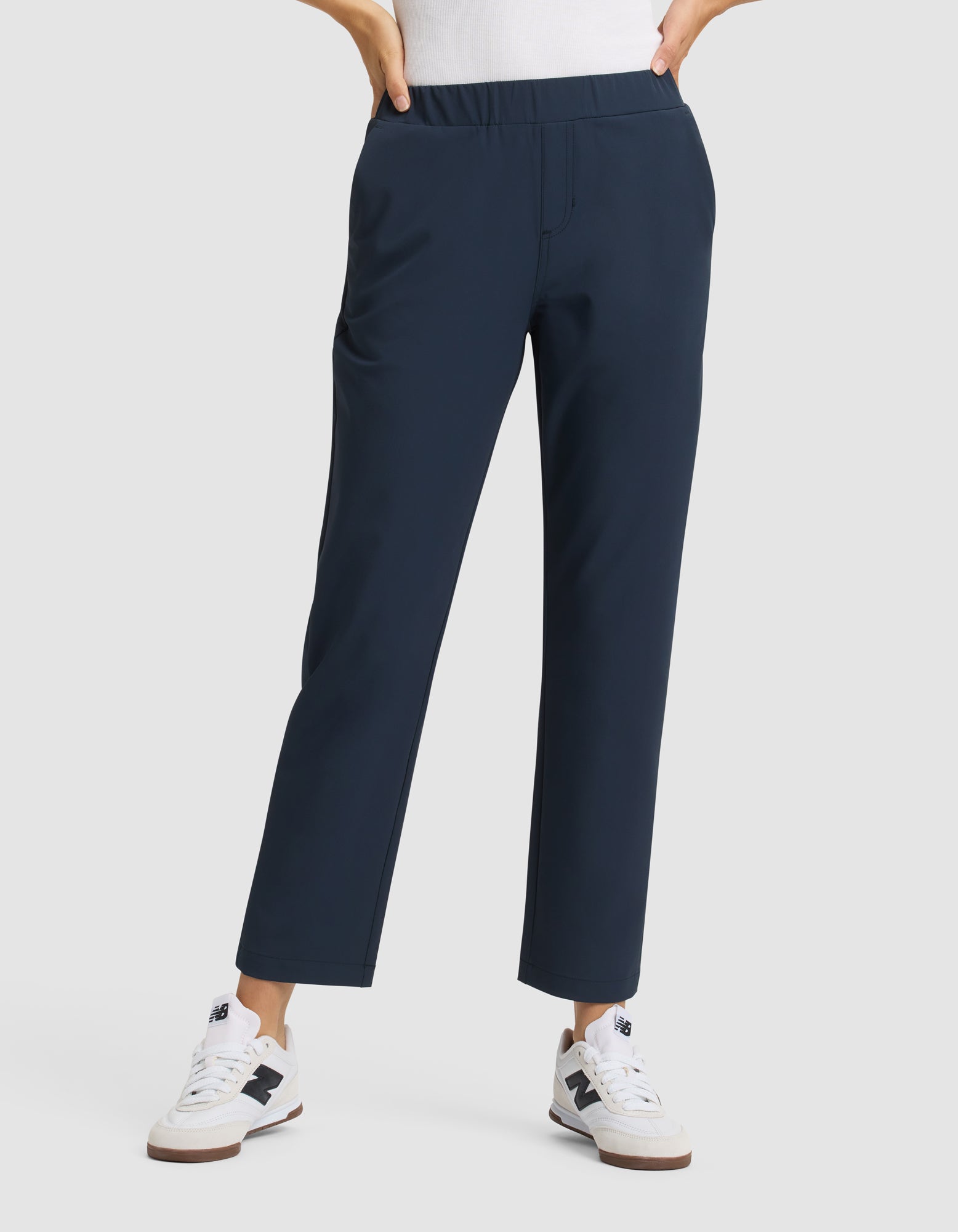 A person stands against a plain light background wearing Cozy Earth's Women's Always Cropped Pant in navy blue with an elastic waistband, paired with a white top and white New Balance sneakers. Their hands are in their pockets. |Color:Eclipse