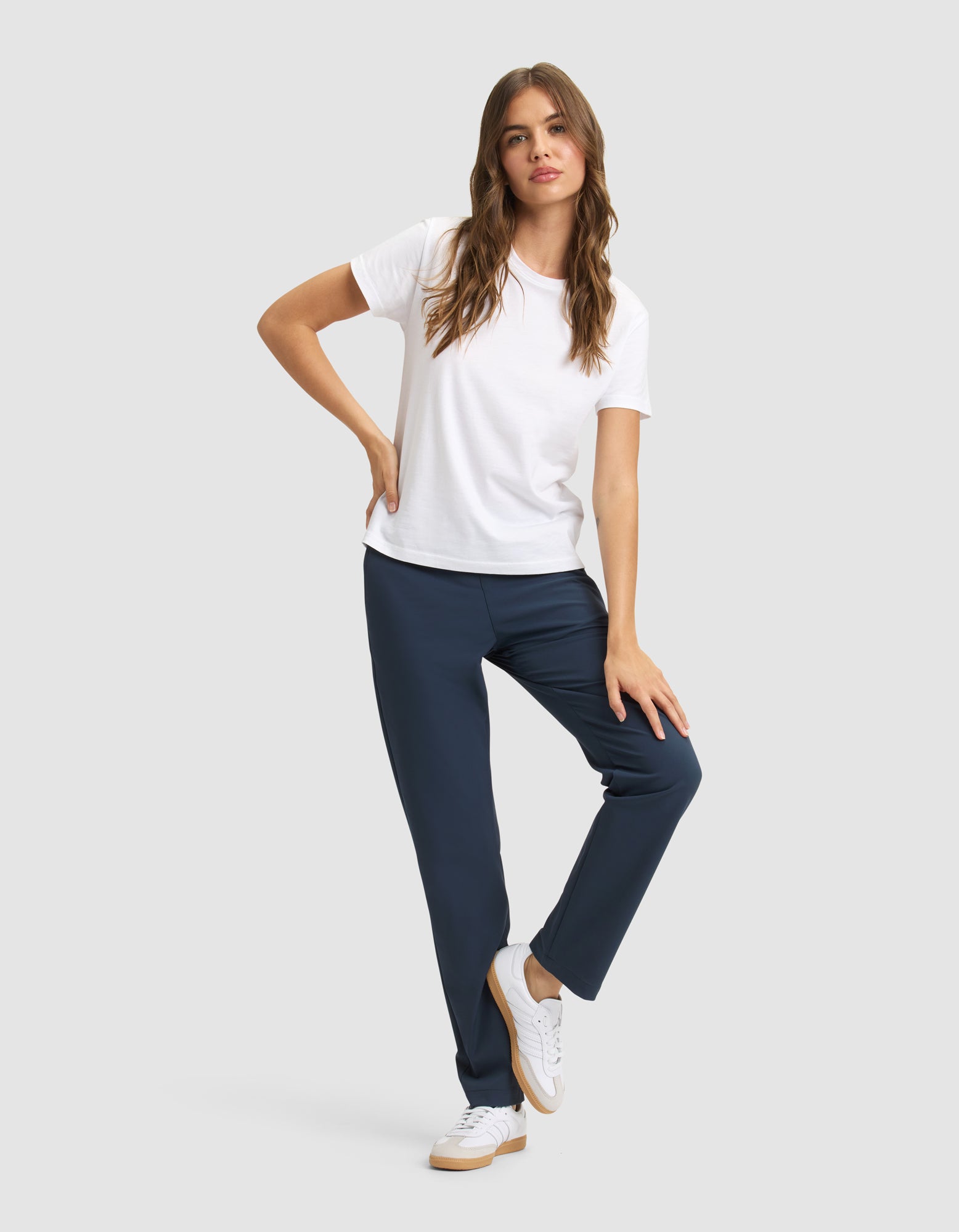 A woman with long, wavy brown hair poses against a plain background, wearing Cozy Earth's Women's Always Pant in navy blue, a white T-shirt, and white sneakers, with one hand on her hip and one leg slightly bent. 
