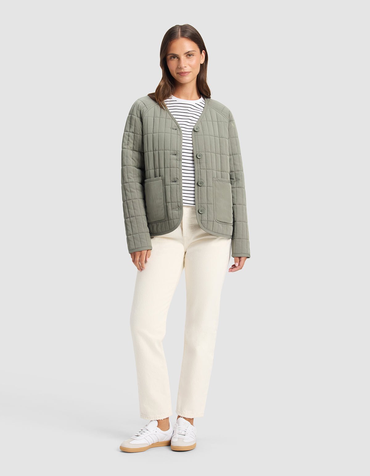 A woman stands facing forward against a light gray background, wearing Cozy Earth's Women's Andie Quilted Jacket in light olive green over a white and black striped shirt, cream pants, and white sneakers.