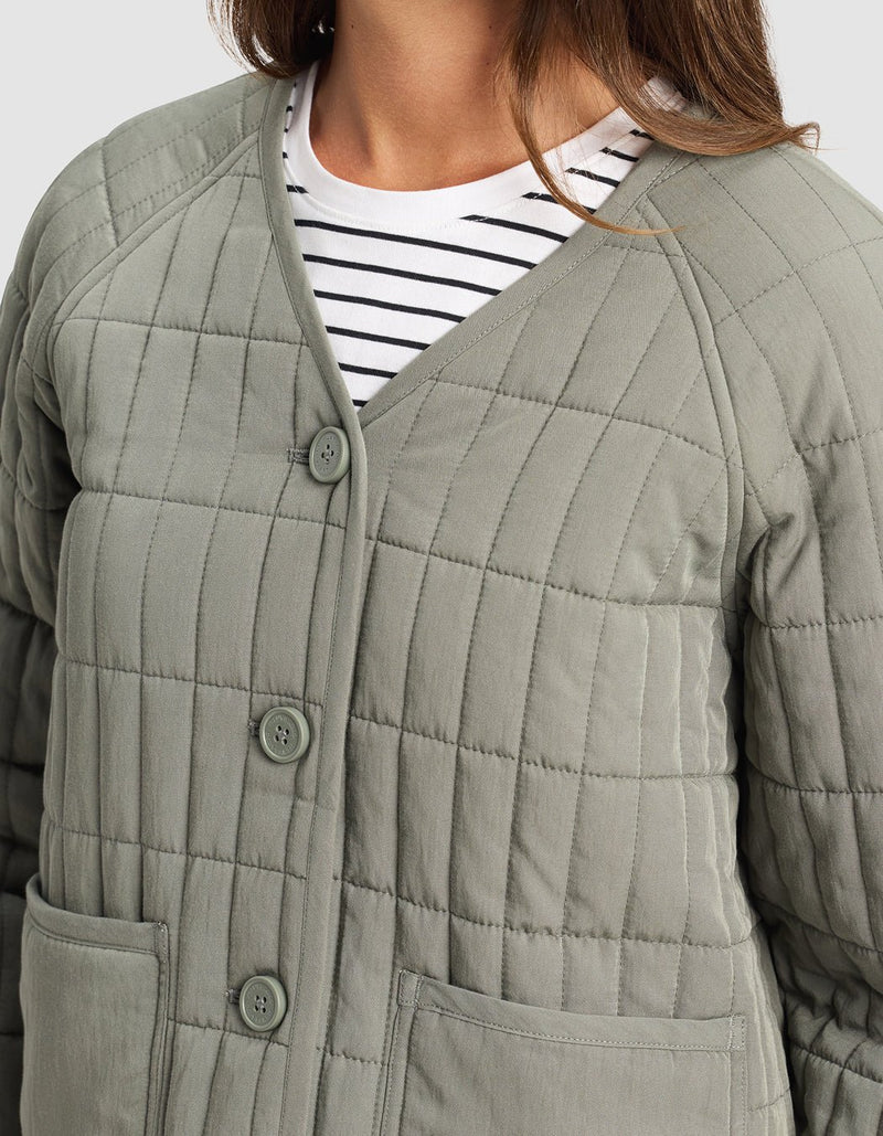 A woman wears the Cozy Earth Women's Andie Quilted Jacket in light gray with large buttons, a V-neck, and two front pockets over a white tee with black stripes. Only her upper body is visible.