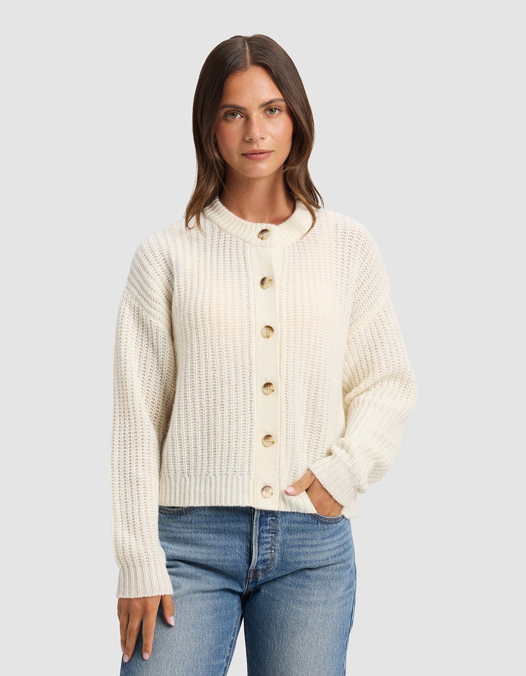 A woman with straight brown hair wears the Cozy Earth Women's Arden Cardigan in cream and blue jeans, standing against a plain light gray background, facing forward with one hand in her pocket.