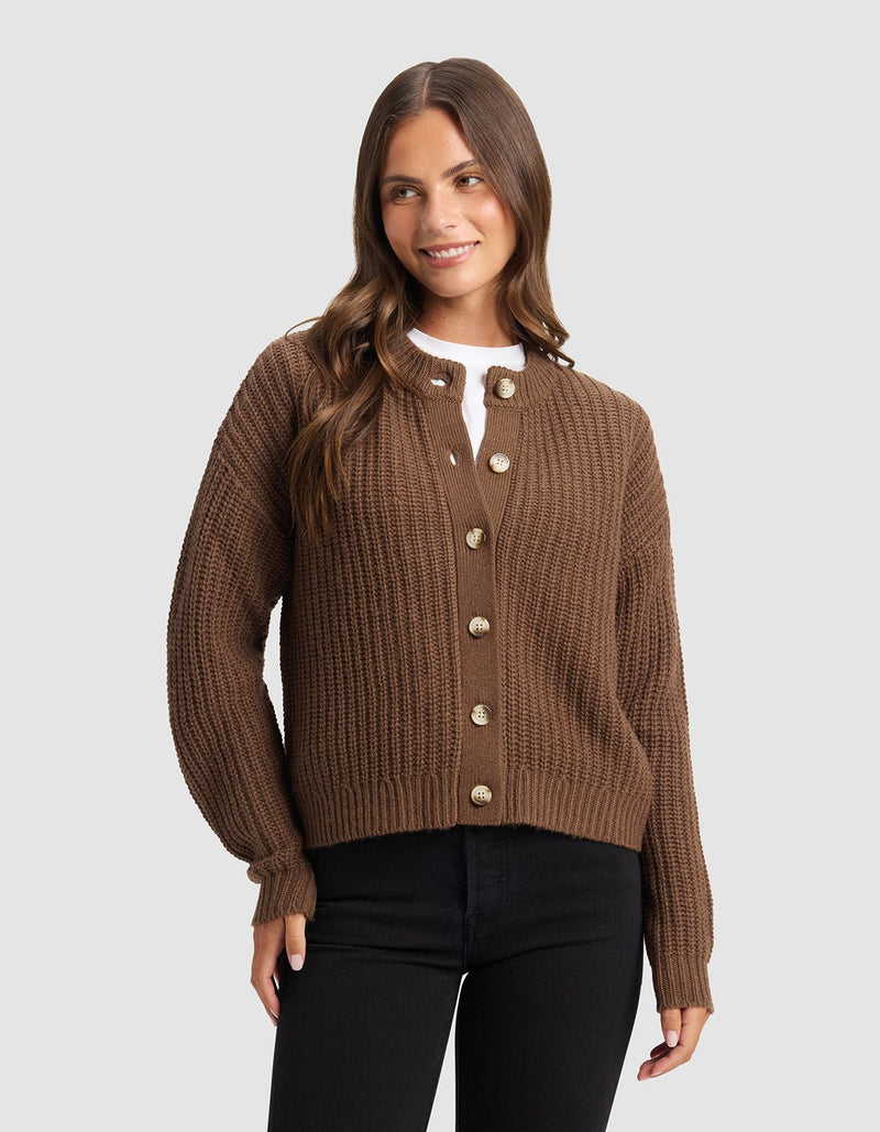 A woman with long brown hair stands smiling against a light gray background, wearing the Cozy Earth Women's Arden Cardigan over a white top and black pants.