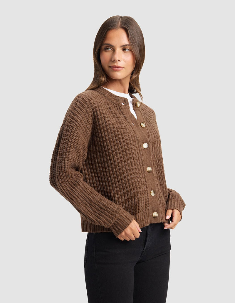A woman with long brown hair models the Cozy Earth Women's Arden Cardigan, a brown ribbed button-up, layered over a white top and black pants against a plain light gray background.