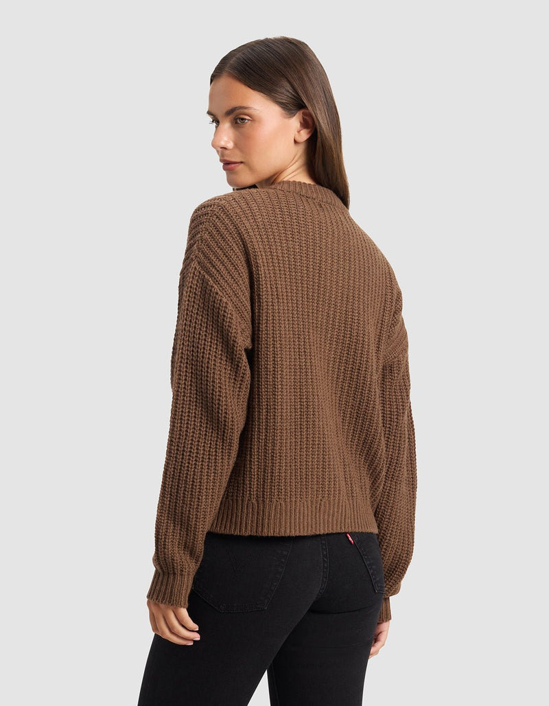 A woman with long brown hair models the Cozy Earth Women's Arden Cardigan, standing against a plain light gray background and looking over her shoulder toward the camera.