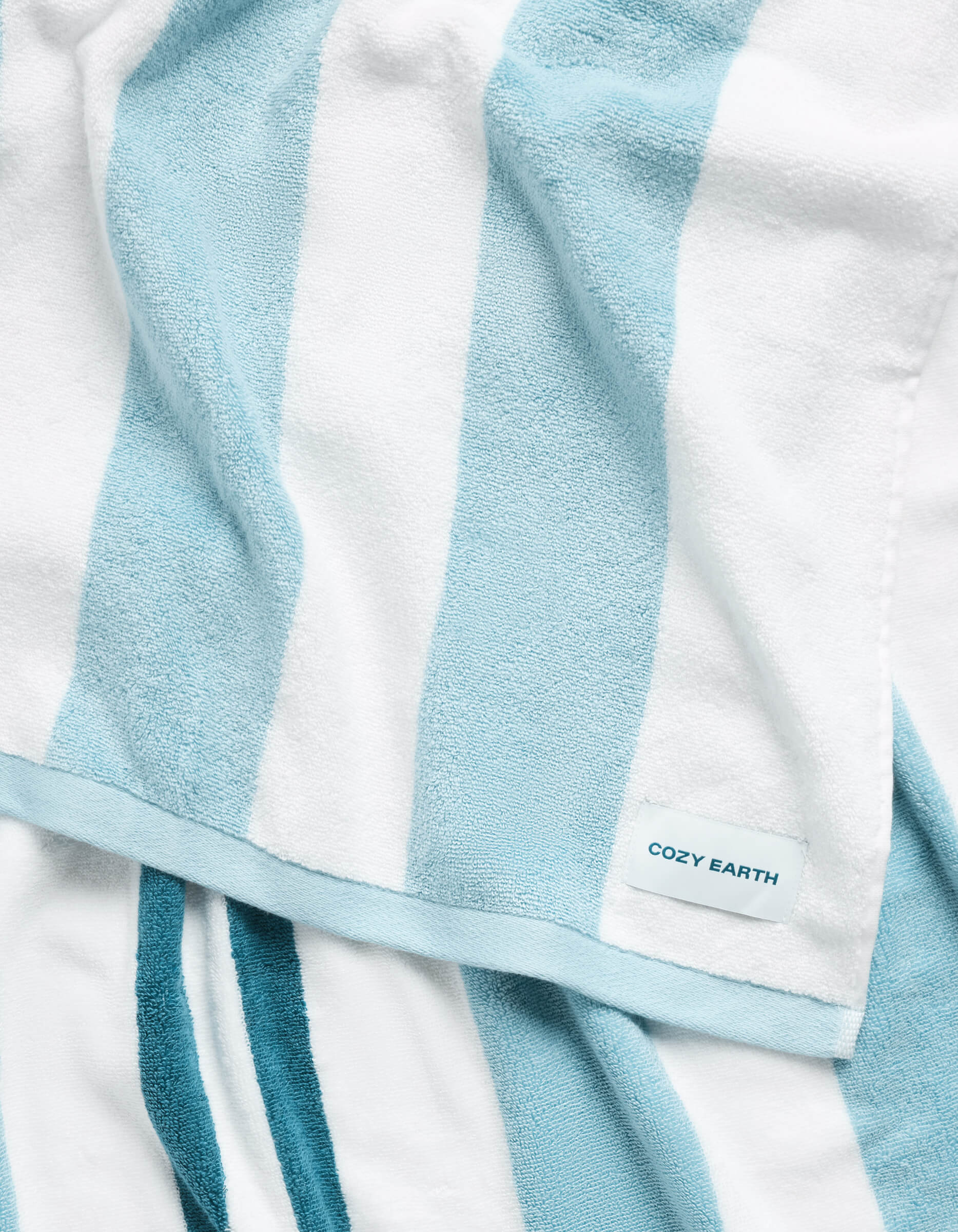 A close-up of the Cozy Earth Classic Stripe Resort Towel, showcasing its soft texture and wide white and light blue vertical stripes, with a small "COZY EARTH" label in the corner.|Color:Wave Stripe