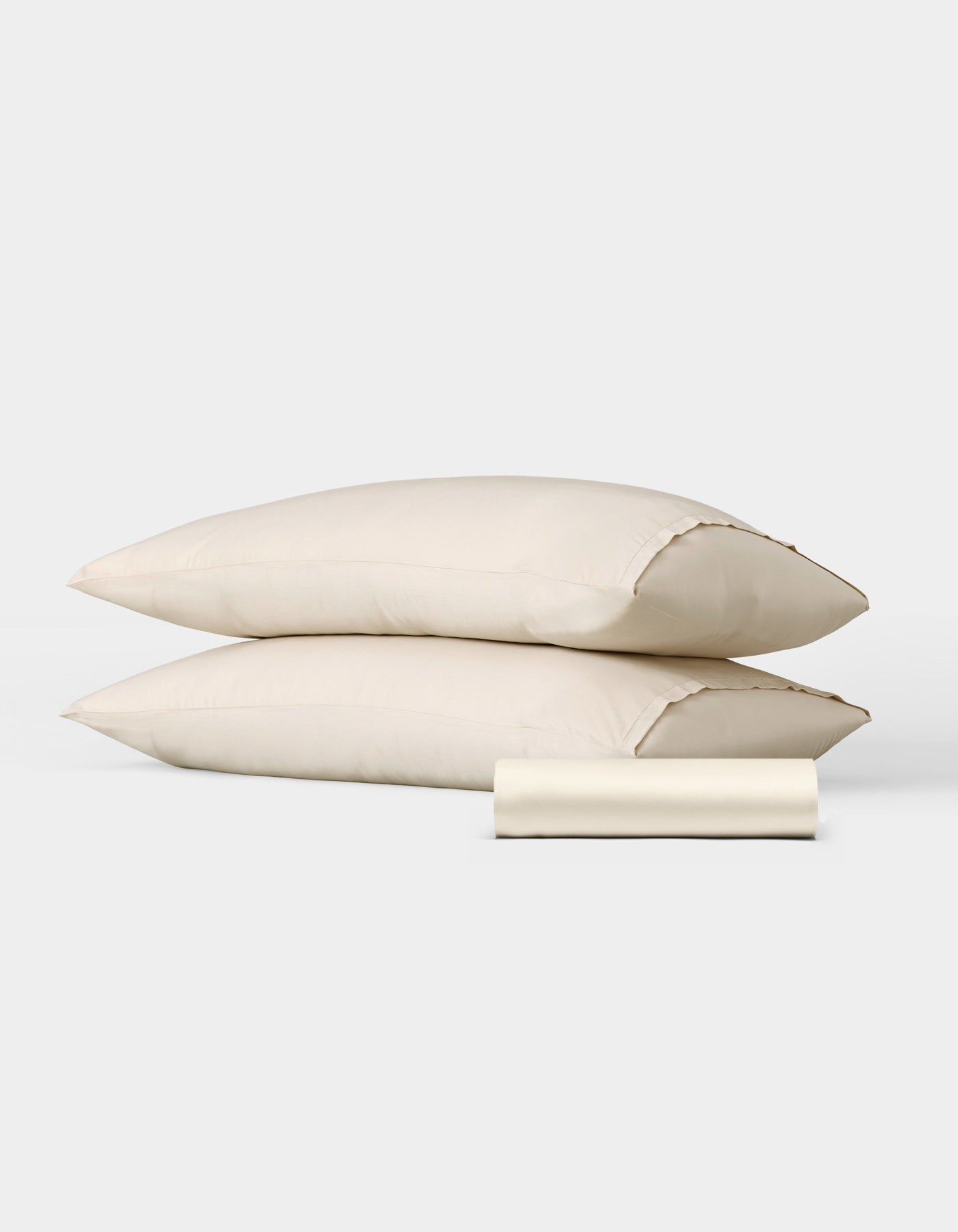 The Bamboo Bedding Basic Bundle by Cozy Earth features two cream-colored pillows stacked together with a matching rolled pillowcase in front, displayed against a plain white background.|Color:Creme