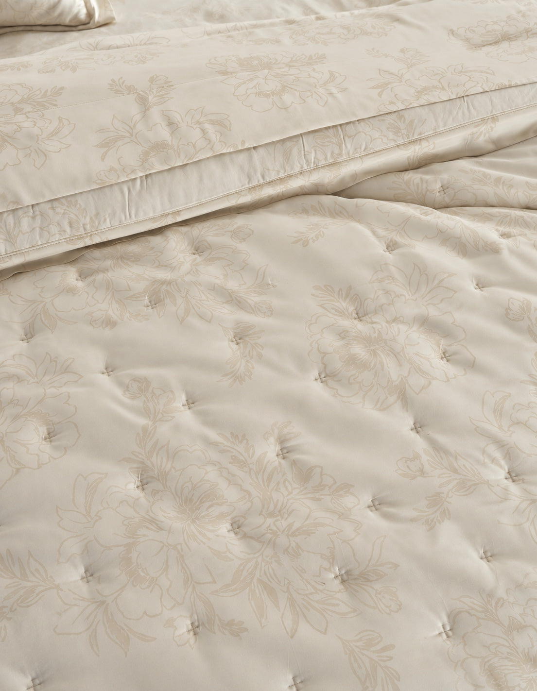 Close-up of a cream-colored quilted bedding set with a subtle floral jacquard pattern, showing the texture of the tufted comforter and matching pillowcase. 