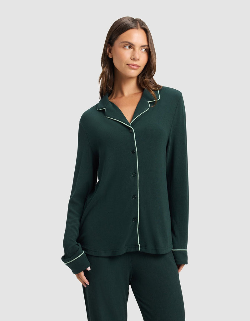 A woman wears the Cozy Earth Women's Bamboo Rib-Knit Classic Long Sleeve Pajama Top, dark green with white piping, standing against a plain light gray background and looking to the side with one hand in her pocket.