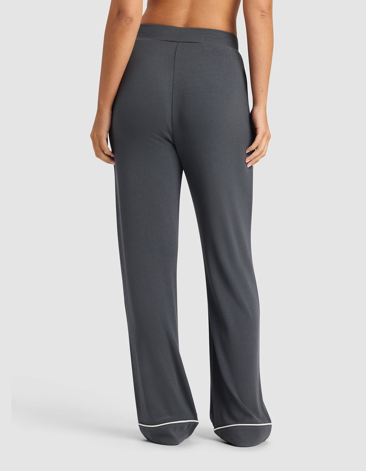 A woman is shown from the back wearing Cozy Earth's Women's Bamboo Rib-Knit Classic Pajama Pant in dark gray with white trim, standing barefoot against a plain light background.|Color:Graphite