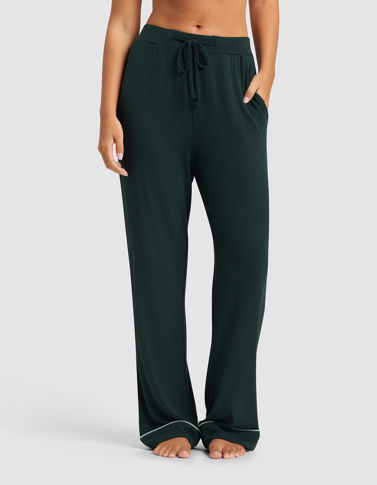A woman wears Cozy Earth's Women's Bamboo Rib-Knit Classic Pajama Pant in dark green, stands barefoot with hands in pockets against a plain light gray background.