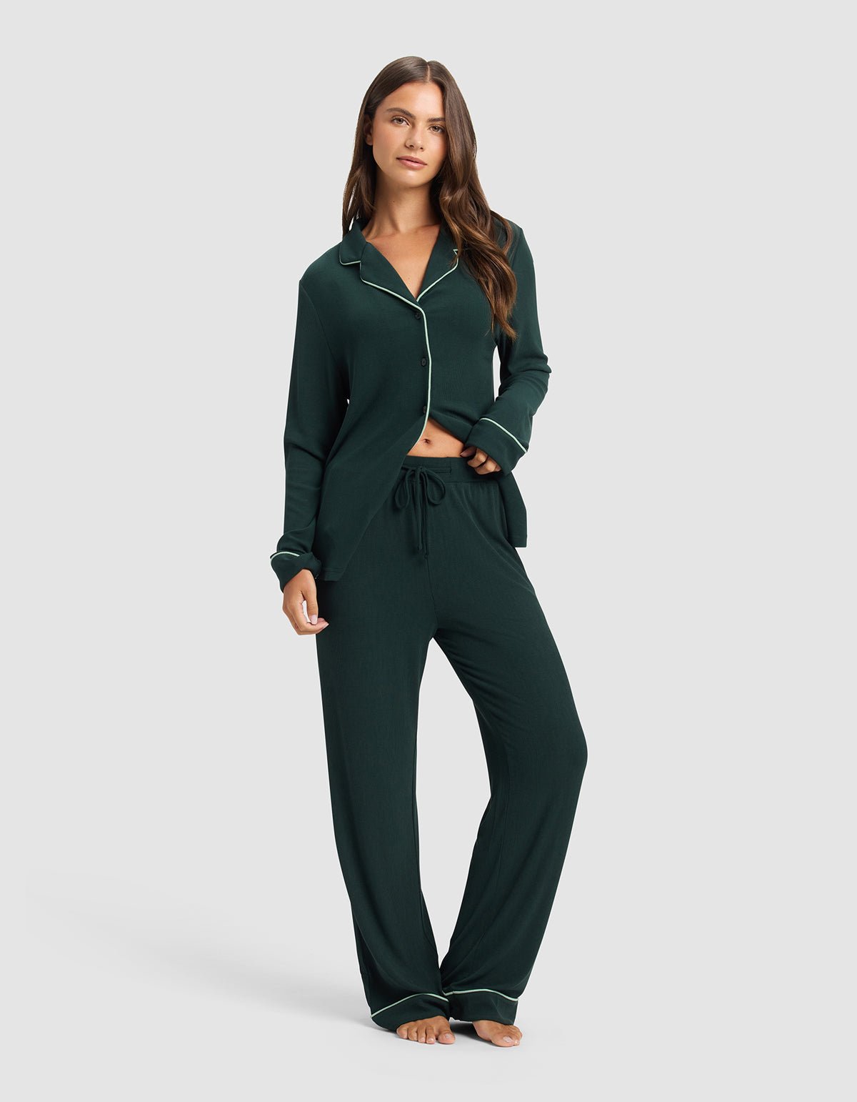 A woman stands barefoot in Cozy Earth's Women's Bamboo Rib-Knit Classic Pajama Pant, dark green with white piping and a drawstring waist. The long-sleeved top is buttoned and slightly open at the bottom. Background: plain light gray.