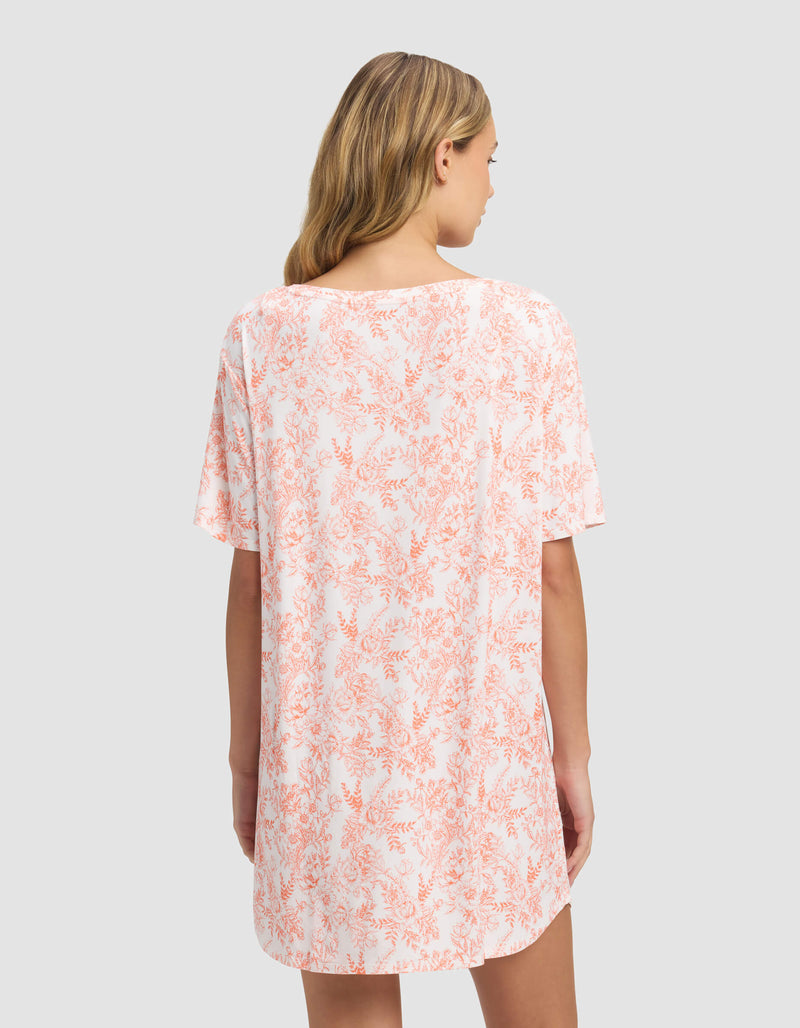 A woman with long blonde hair is seen from the back wearing the Cozy Earth Women’s Bamboo Stretch-Knit Sleep Dress, a loose short-sleeved outfit in a light pink and white floral pattern, set against a plain gray background.