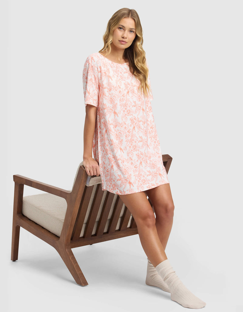 A woman in the Cozy Earth Women's Bamboo Stretch-Knit Sleep Dress, featuring loose short sleeves and a pink floral print, with cream socks, leans against a wooden chair with a neutral cushion against a light gray background.