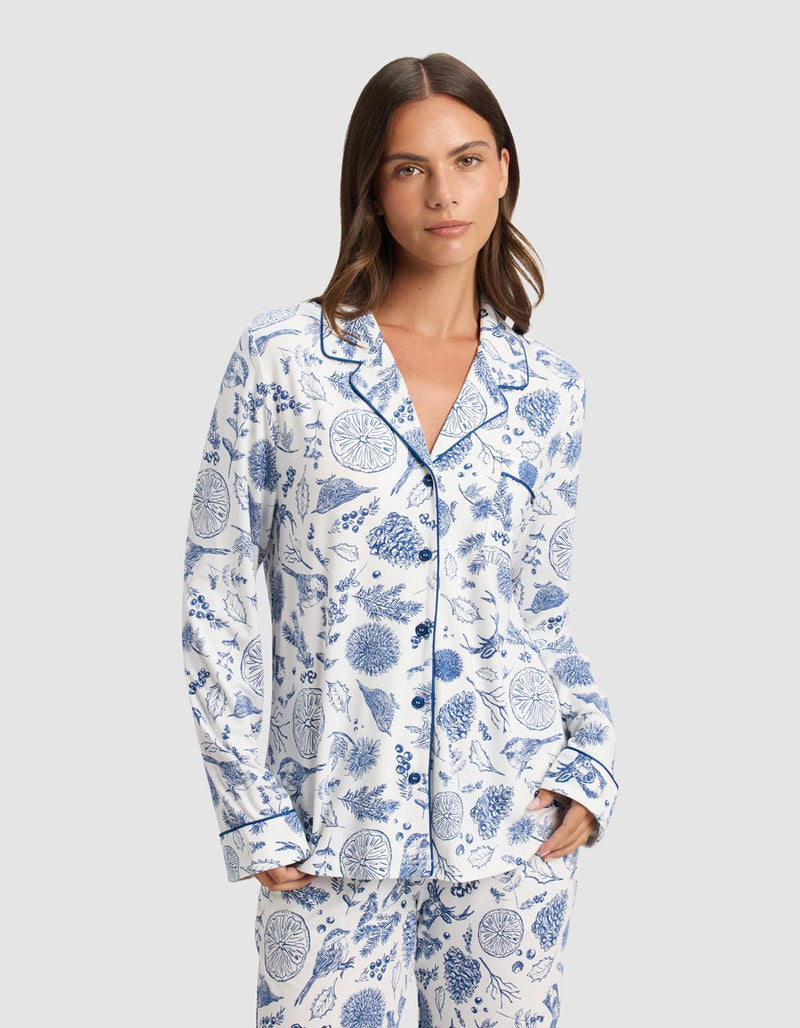 A woman with straight brown hair wears the Cozy Earth Women's Bamboo Stretch-Knit Long Sleeve Pajama Top, featuring a notched collar and blue botanical prints, standing with her hands in her pockets against a plain light background.
