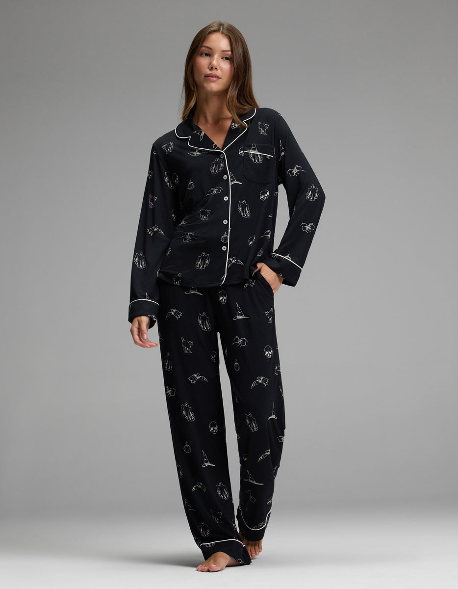 A woman stands barefoot in a relaxed pose, wearing the Cozy Earth Women’s Bamboo Stretch-Knit Long Sleeve Pajama Set—black with white piping and small white illustrated patterns—against a plain gray background.|Color:Halloween Print