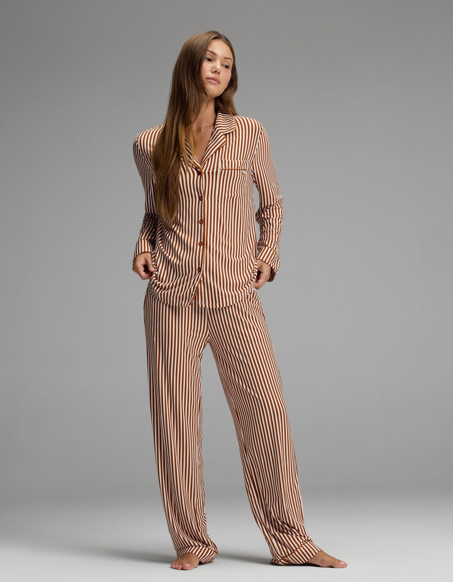 A person stands barefoot on a gray floor, wearing the Cozy Earth Women’s Bamboo Stretch-Knit Long Sleeve Pajama Set in brown-and-white vertical stripes, featuring loose-fitting long sleeves and pants, against a plain gray background.|Color:Market Stripe in Cinnamon