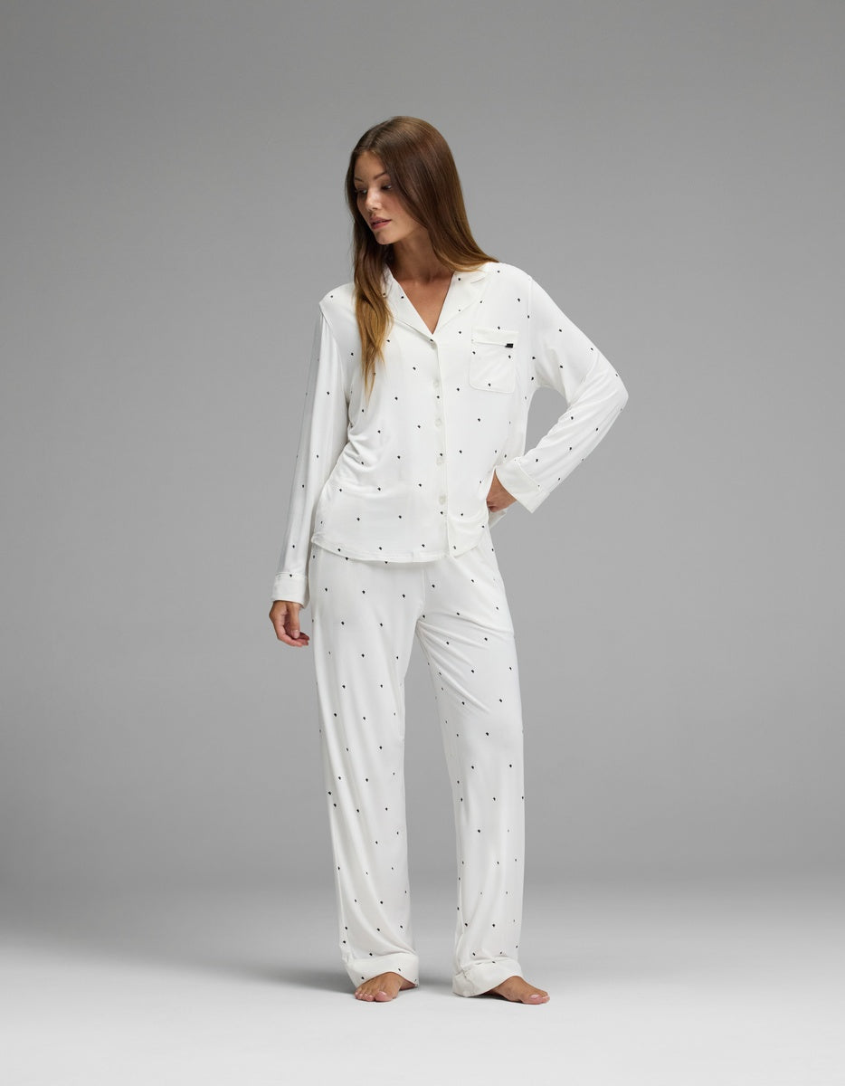 A woman models the Cozy Earth Women’s Bamboo Stretch-Knit Long Sleeve Pajama Set in white with small black polka dots, standing barefoot on a gray background and looking to her left with one hand on her hip.|Color:Ivory Mini Ghost