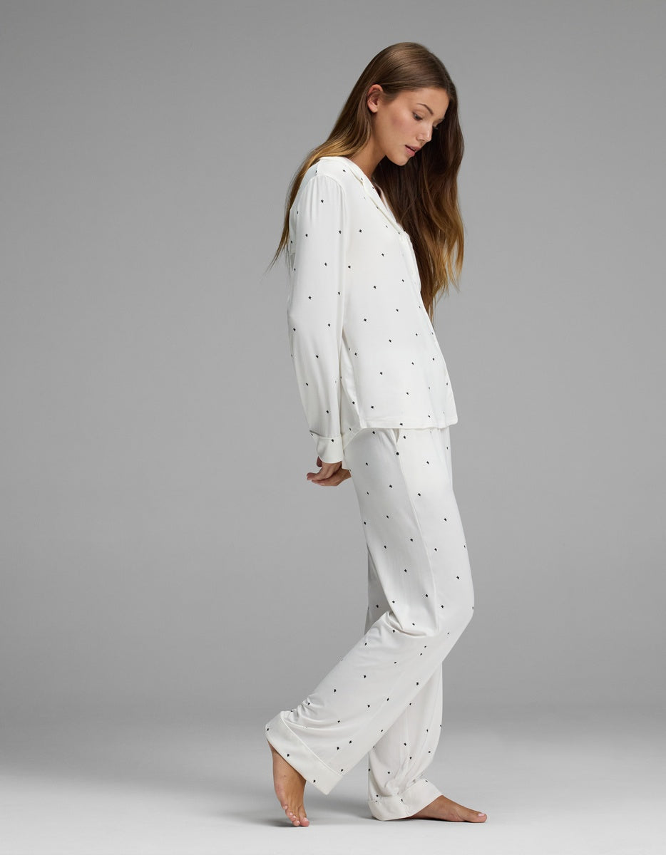 A woman models the Cozy Earth Women’s Bamboo Stretch-Knit Long Sleeve Pajama Set in white with small black dots, standing barefoot on a gray background in a side profile with her head gently tilted down and her long hair draped over one shoulder.|Color:Ivory Mini Ghost