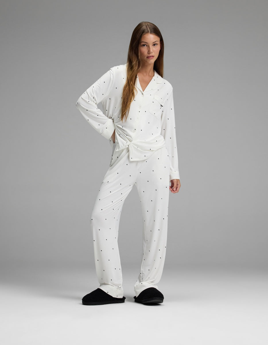 A woman wears the Cozy Earth Women’s Bamboo Stretch-Knit Long Sleeve Pajama Set in white polka dot print, paired with black slippers, as she stands in a relaxed pose against a plain gray background.|Color:Ivory Mini Ghost