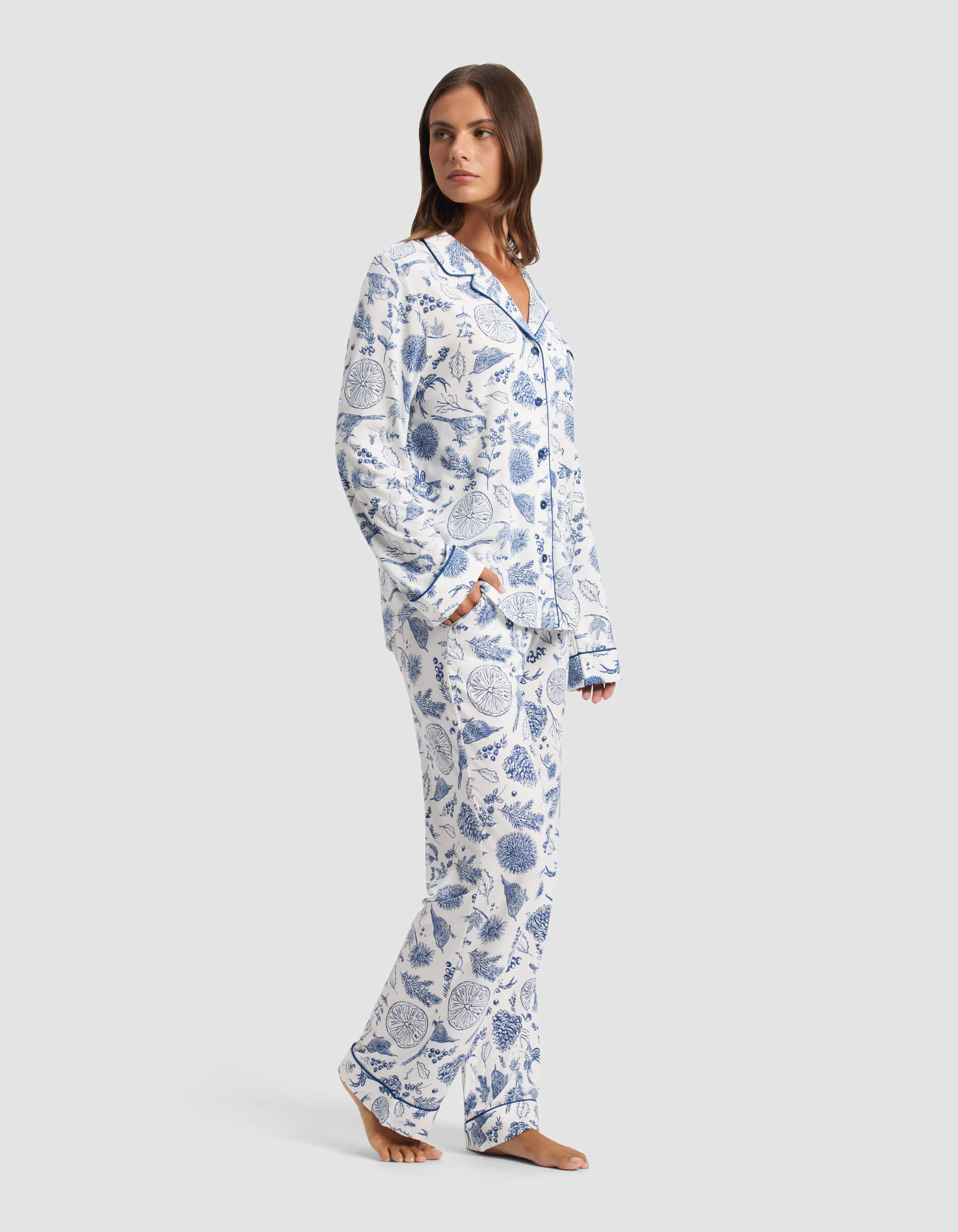 A woman stands barefoot wearing Cozy Earth's Women's Bamboo Stretch-Knit Pajama Pant, featuring a white background with blue botanical print and paired with a matching long-sleeve button-up top. She looks over her shoulder. |Color:Winter Toile Pacific