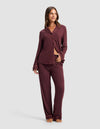 A woman stands barefoot, smiling with one hand on her waist, wearing the Cozy Earth Women's Bamboo Stretch-Knit Long Sleeve Pajama Set—a matching maroon outfit featuring a collared, button-up top with a chest pocket and matching pants. |Color:Burgundy