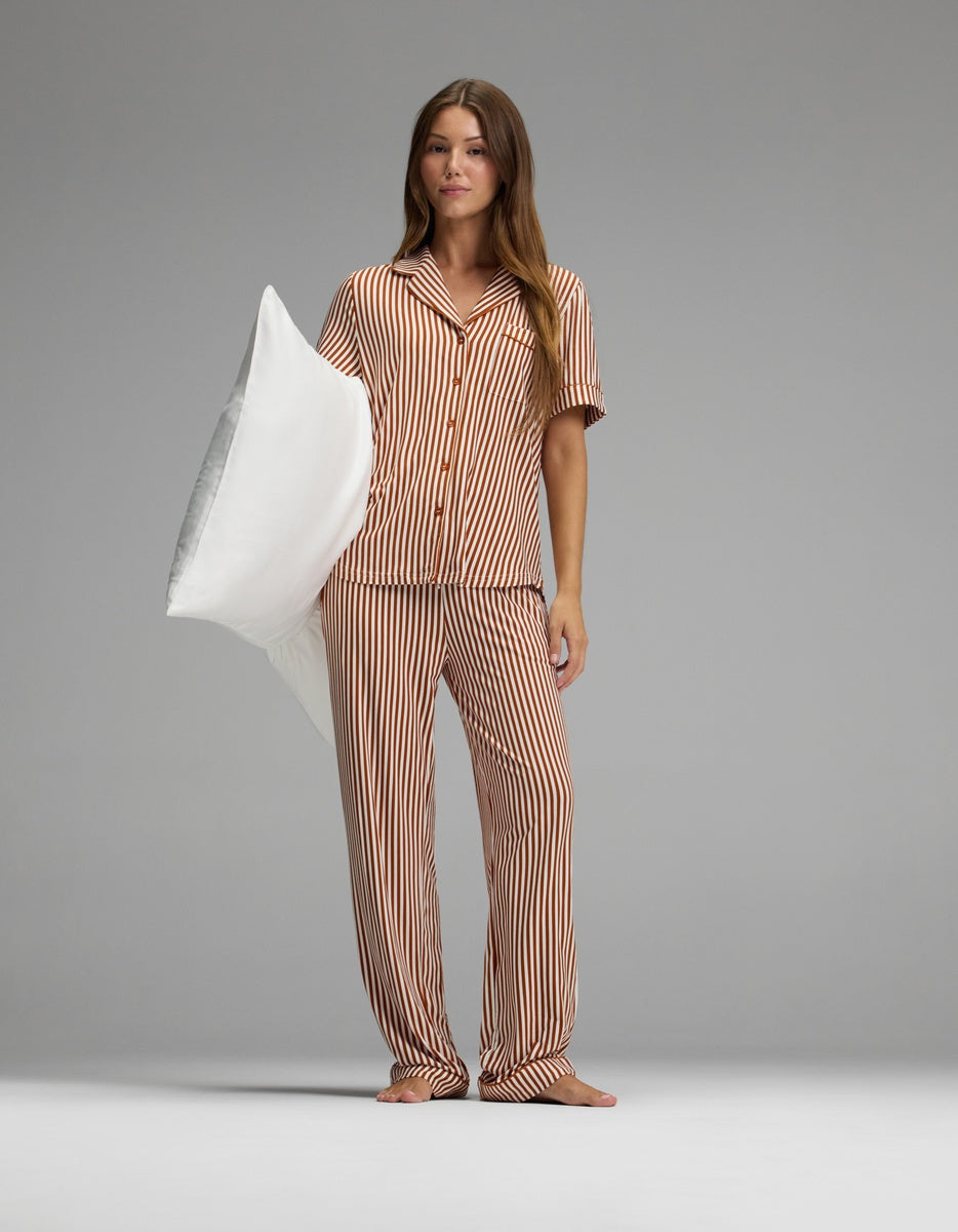 A woman stands barefoot on a gray floor, wearing Cozy Earth’s Women’s Bamboo Stretch-Knit Pajama Pant in an orange-and-white striped pattern and holding a large white pillow under her left arm. The background is plain and gray with evenly soft lighting.|Color:Market Stripe in Cinnamon