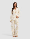 A woman with long brown hair stands barefoot against a light gray background, wearing Cozy Earth’s Women's Bamboo Stretch-Knit Pajama Pant in cream with a gold floral pattern, looking forward with a relaxed expression.|Color:Winter Toile Dijon