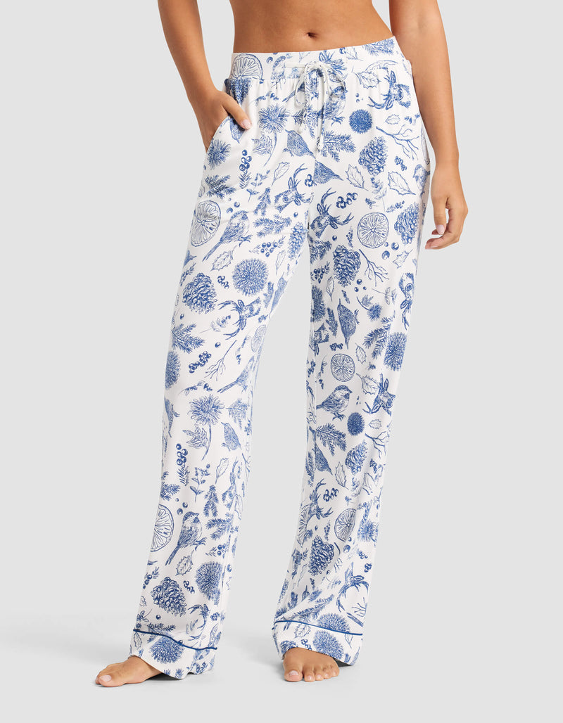 A woman stands barefoot against a plain background, wearing Cozy Earth's Women's Bamboo Stretch-Knit Pajama Pant—loose-fitting white pants with a blue botanical print and a drawstring waist.