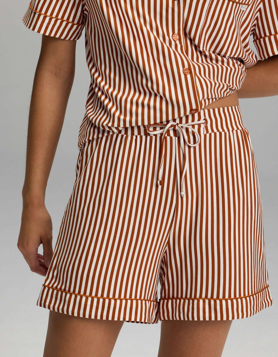 A person is shown from shoulder to mid-thigh wearing the Cozy Earth Women’s Bamboo Stretch-Knit Pajama Short, part of a matching short-sleeve button-up shirt and shorts set featuring vertical white and brown stripes.|Color:Market Stripe in Cinnamon