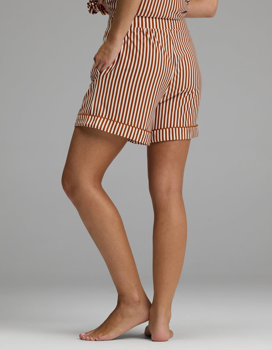 A person standing barefoot on one leg wears Cozy Earth Women’s Bamboo Stretch-Knit Pajama Shorts in a brown and white vertical stripe pattern, with their upper body mostly out of frame against a plain gray background.|Color:Market Stripe in Cinnamon