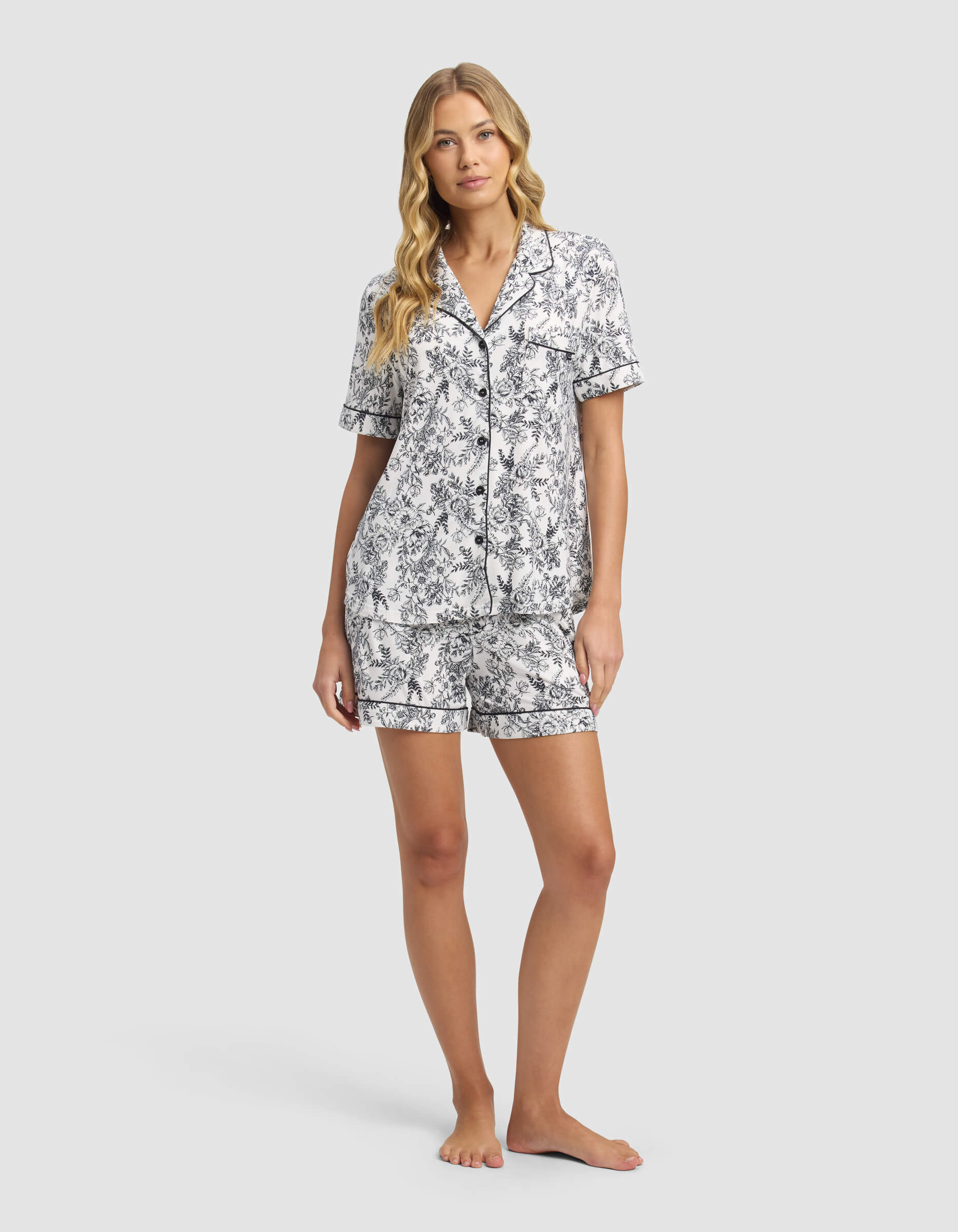 A woman with long, wavy blonde hair poses barefoot against a light gray background, wearing Cozy Earth’s Women's Bamboo Stretch-Knit Short Sleeve Pajama Set in black and white floral print shorts and matching button-up top.|Color:Jet Black Toile