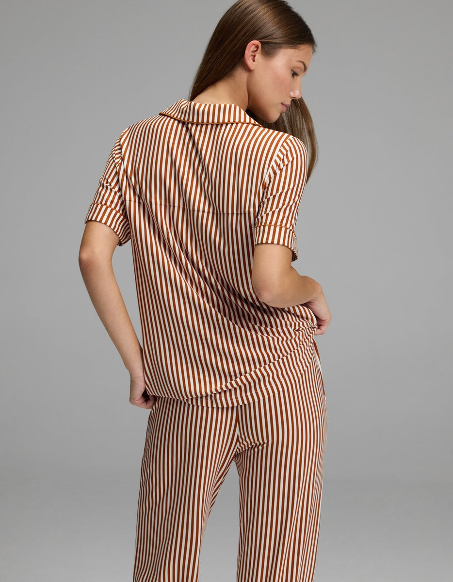 A person stands with their back to the camera, wearing the Cozy Earth Women’s Bamboo Stretch-Knit Short Sleeve Pajama Top paired with matching pants featuring vertical brown and white stripes. The background is plain gray.|Color:Market Stripe in Cinnamon