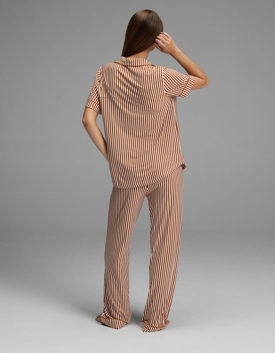 A person stands facing away, wearing the Cozy Earth Women’s Bamboo Stretch-Knit Short Sleeve & Pant Pajama Set in a loose fit with tan and white vertical stripes. They have long, straight brown hair and hold one arm bent at the elbow, touching the side of their head. The background is plain gray.|Color:Market Stripe in Cinnamon