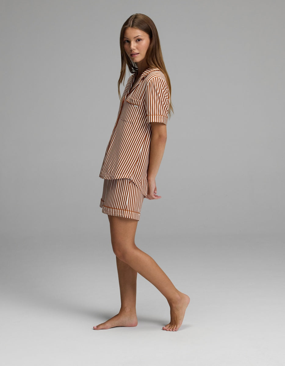 A woman stands barefoot on a gray background, wearing the Cozy Earth Women’s Bamboo Stretch-Knit Short Sleeve Pajama Set in a brown-and-white striped pattern with shorts. She is turned slightly to the side and looking toward the camera.|Color:Market Stripe in Cinnamon