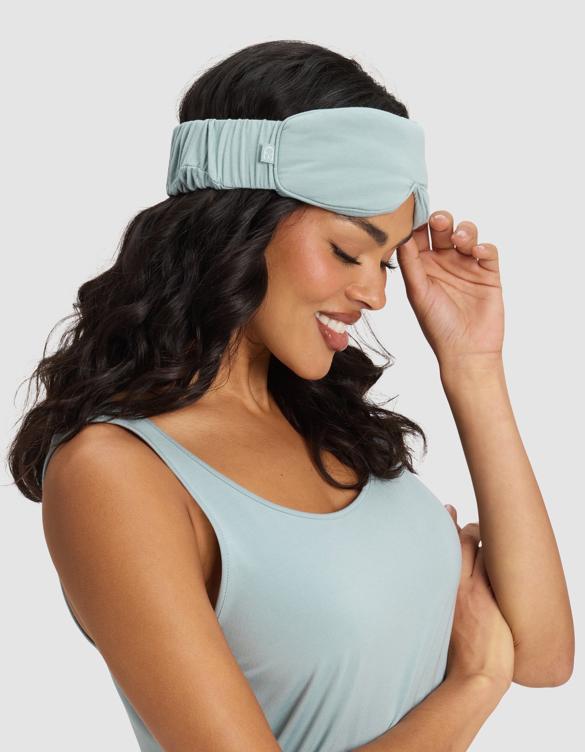 A woman with wavy dark hair smiles while wearing a Cozy Earth Bamboo Stretch-Knit Sleep Mask in light blue and a matching tank top, gently touching the mask with one hand. The background is plain light gray. 