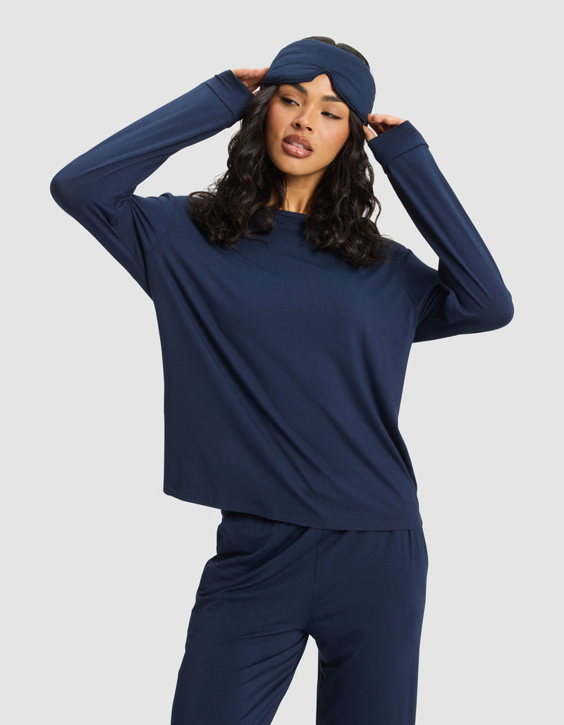 A woman wearing a navy blue pajama set poses with her hands touching the sides of a Cozy Earth Bamboo Stretch-Knit Sleep Mask, standing against a plain light background.