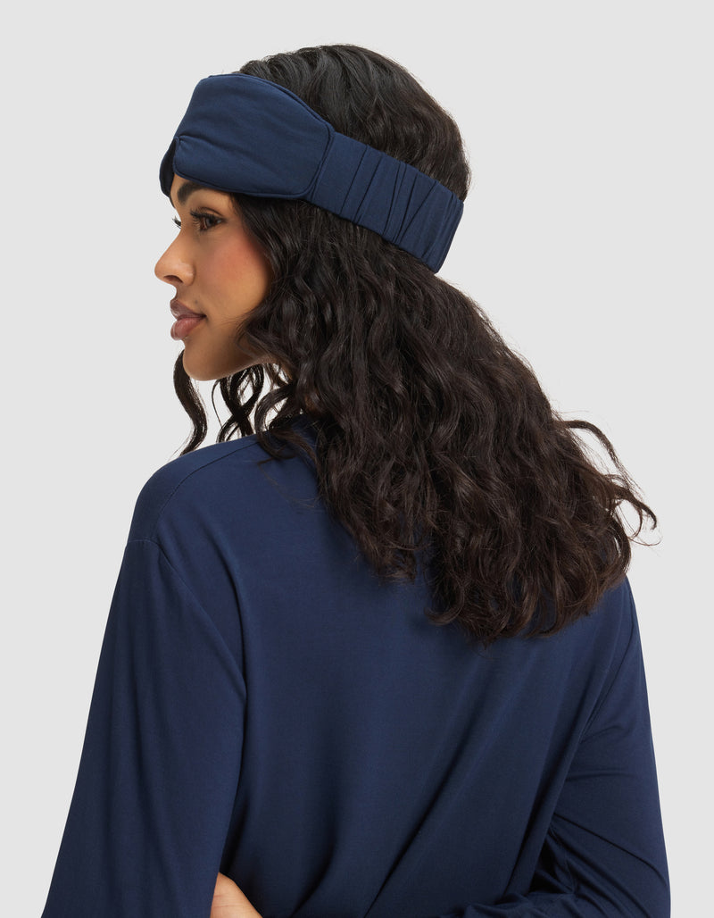 A woman with long, wavy dark hair is shown from the side and back wearing the Cozy Earth Bamboo Stretch-Knit Sleep Mask in navy blue, paired with a matching top against a plain light gray background.
