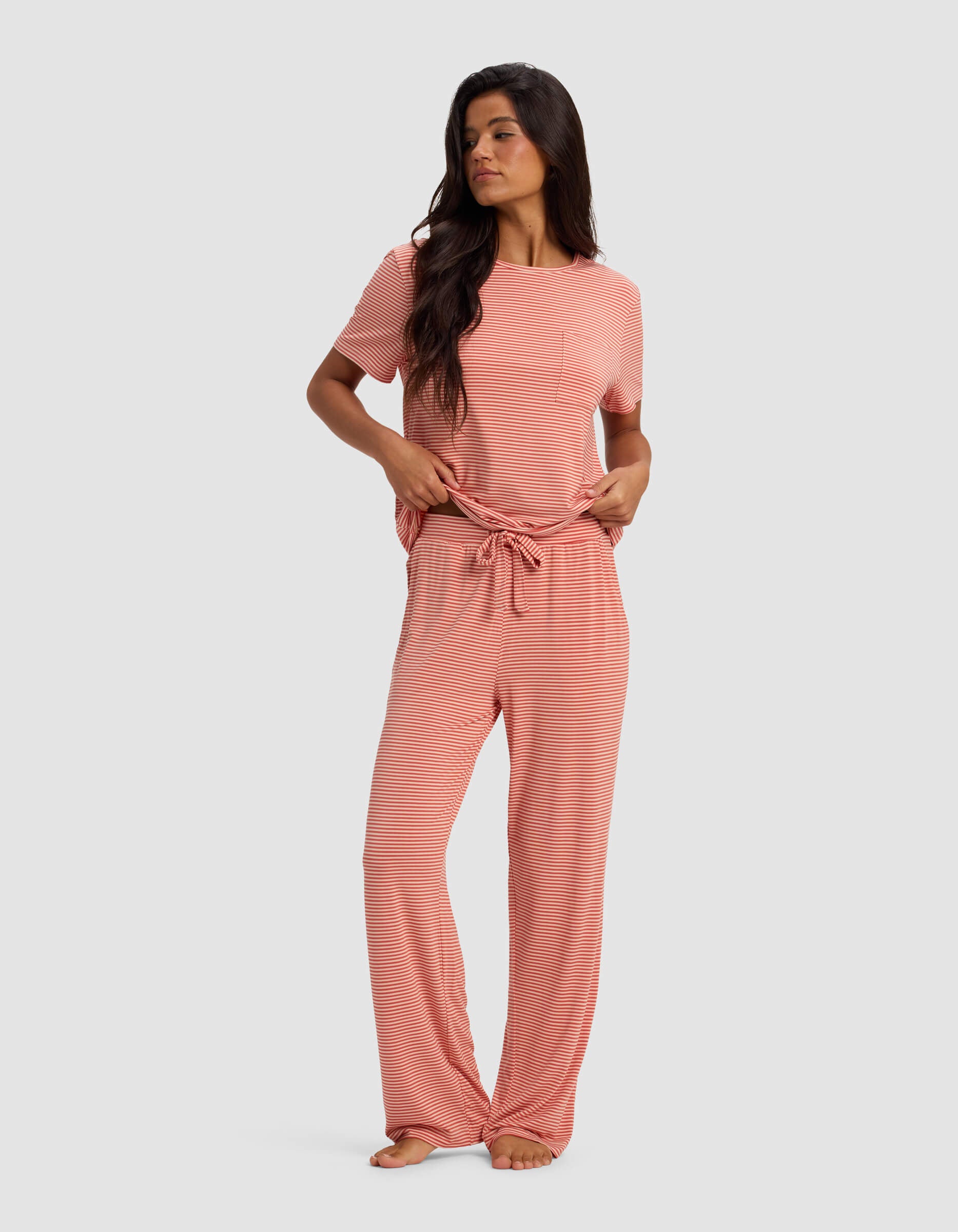 A woman in the Cozy Earth Women's Bamboo Stretch-Knit Lounge Tee & Pant Set, featuring pink and white stripes, short sleeves, front pocket, and drawstring waist, stands barefoot against a plain background, looking to the side.|Color:Sunset Mini Stripe
