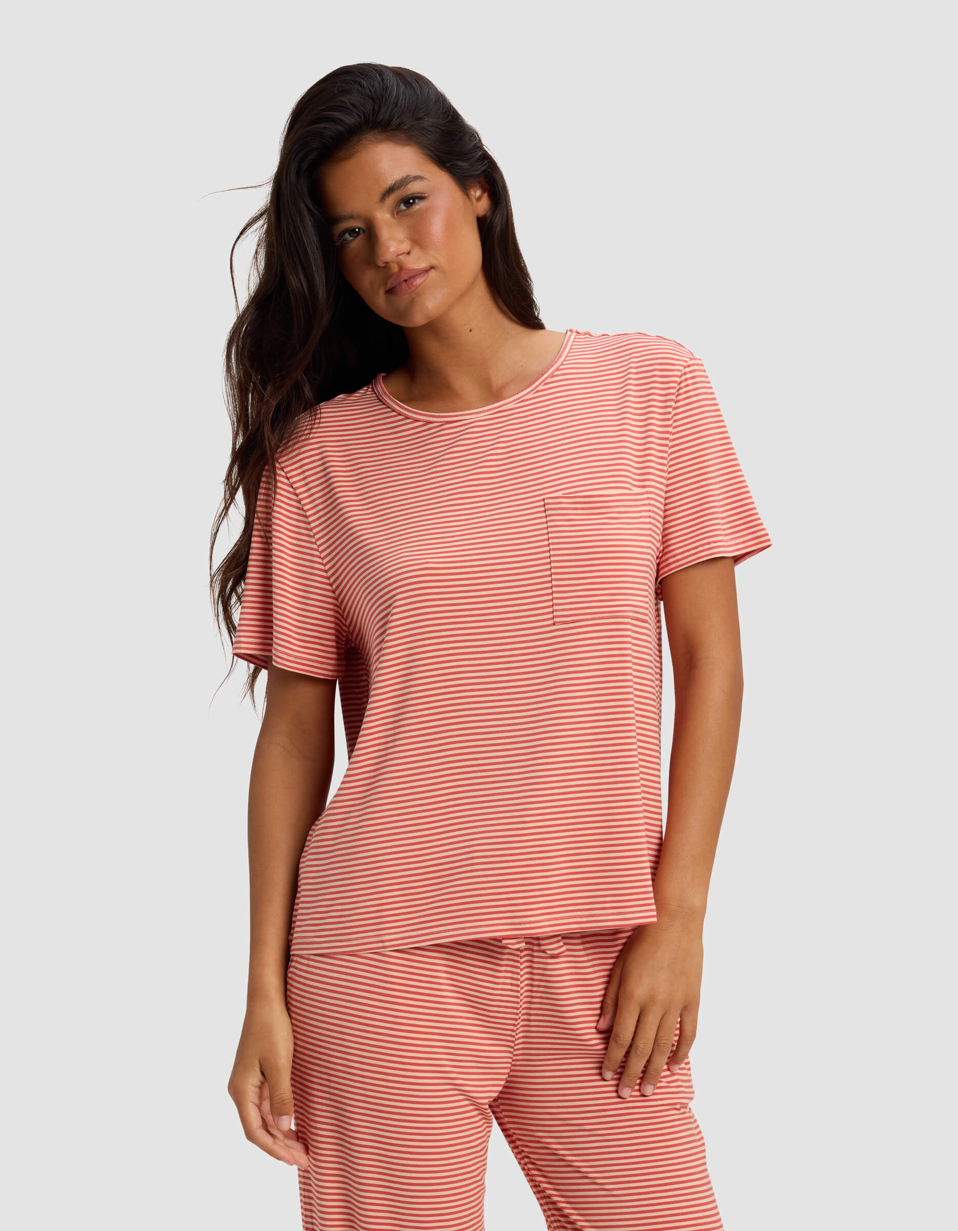 A woman with long dark hair wears the Cozy Earth Women's Bamboo Stretch-Knit Lounge Tee in coral and white stripes, featuring a small chest pocket, while standing against a plain light gray background.|Color:Sunset Mini Stripe
