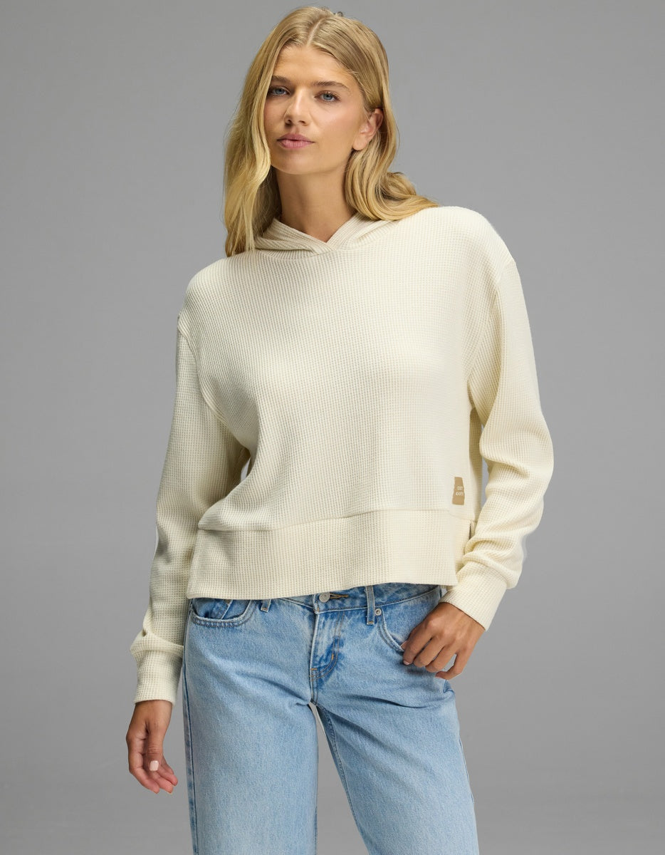 A person wearing a cream-colored Women’s Blake Waffle Hoodie with a small rectangular tag on the side, paired with light-wash blue jeans.|Color:Coconut