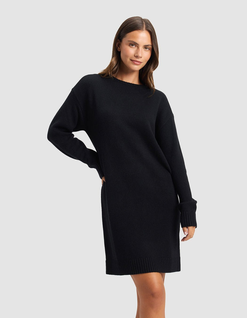 A woman poses against a light gray background, wearing the Cozy Earth Women's Brooklyn Sweater Dress—a loose-fitting, long-sleeved black knit style. She smiles softly with one hand on her hip.