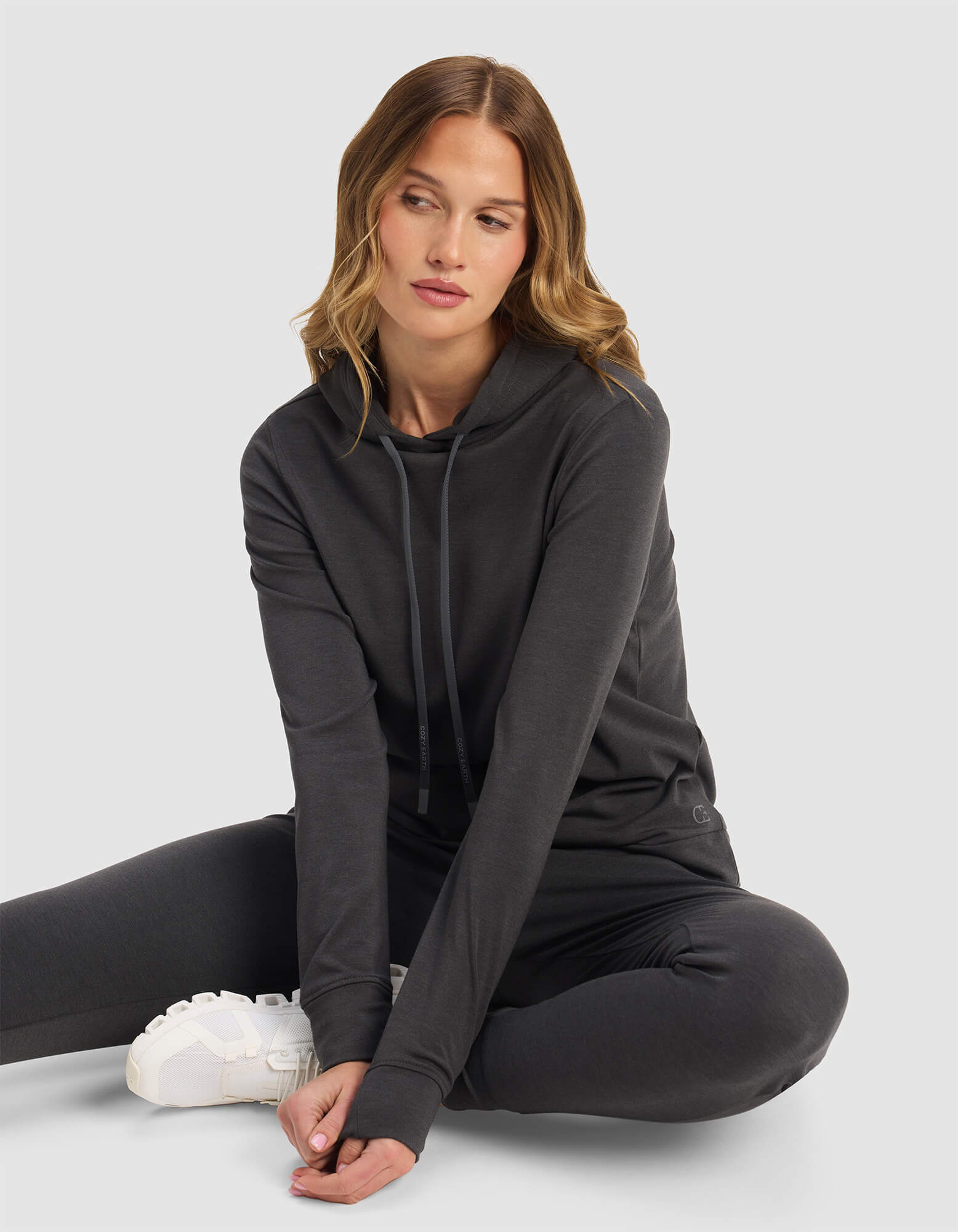 A woman with wavy, light brown hair sits on the floor in a relaxed pose, wearing Cozy Earth's Women's Brushed Bamboo Hoodie in dark gray with matching pants and white sneakers, set against a plain light gray background.|Color:Charcoal